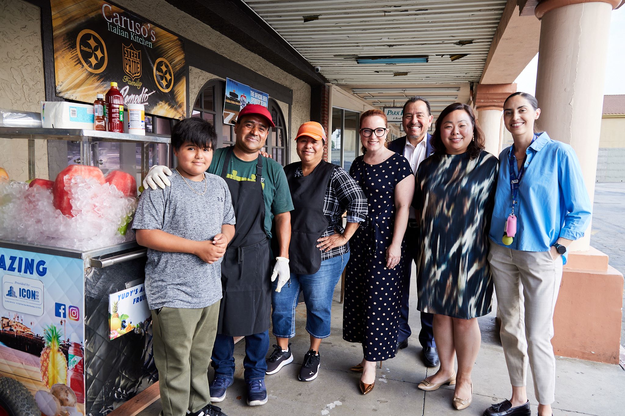 Group photo on a sidewalk outside a storefront: a boy and two vendors in aprons stand beside a fruit cart packed with ice and watermelon, joined by several smiling visitors under a covered walkway.