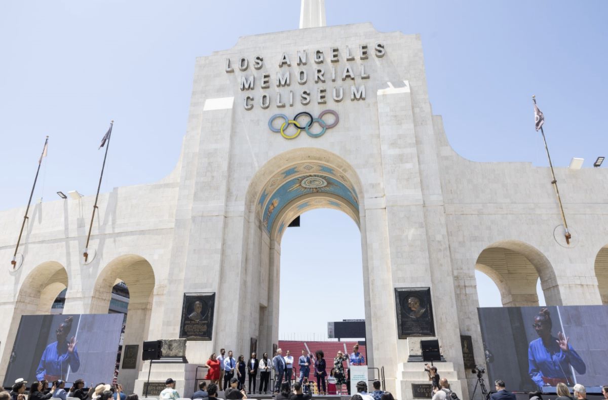 Public event beneath the Los Angeles Memorial Coliseum arch with Olympic rings; speakers on stage and two large video screens.
