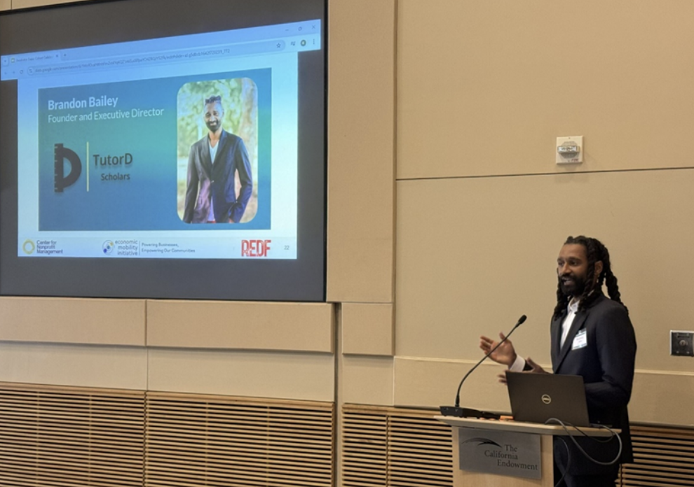 Presenter at a podium beside a projected slide for TutorD Scholars showing a headshot and the title &lsquo;Founder and Executive Director.